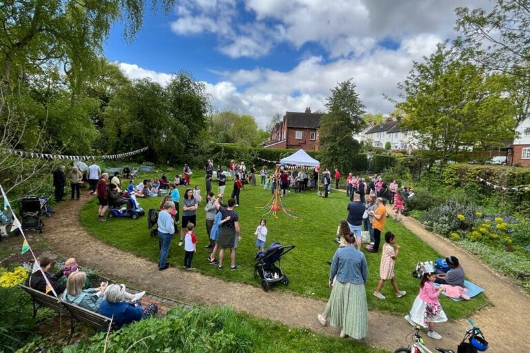 A garden full of people. There is bunting and maypole in the centre of the lawn