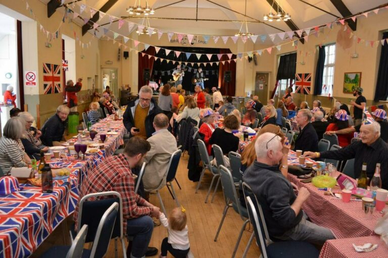 People sat on long tables in Moor Pool Hall. There are Union Flag tablecloths and a band playing on the stage