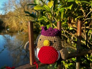 A crochet robin wearing a purple crochet crown sat on a railing in front of a pond