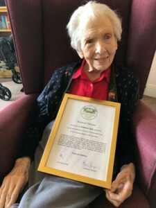 An older woman sitting in an armchair and holding a framed certificate