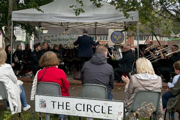 A brass band playing under a gazebo with spectators sat on chairs.